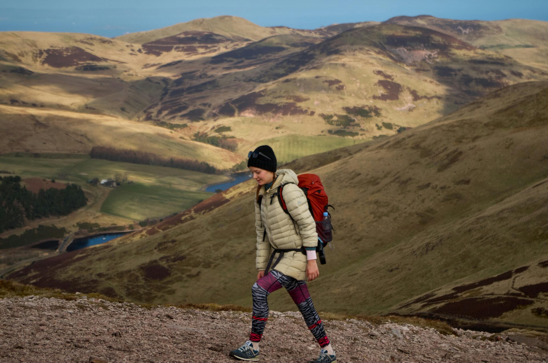 Young female hiker with a red backpack ascending a rocky fell in the Scottish hills, demonstrating what to take on a day hike in high winds and cool weather.